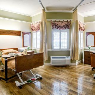 A clean and spacious hospital room with two patient beds, separated by curtains, featuring large windows for natural light.