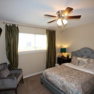 A cozy bedroom featuring a bed with patterned bedding, a chair, curtains, and a ceiling fan.