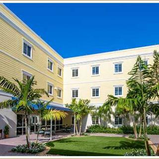 A bright courtyard featuring palm trees and a well-maintained garden, surrounded by a yellow multi-story building under a clear blue sky.