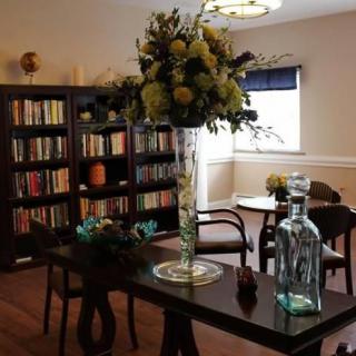 A cozy library corner with bookshelves, a large vase of flowers on a table, and chairs arranged for reading.