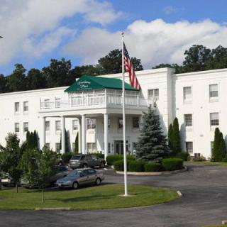 A white hotel building with a green roof, surrounded by trees, with cars parked in front and an American flag displayed.