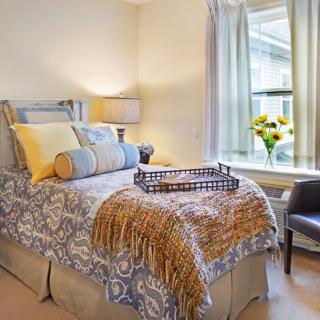 A cozy bedroom with an elegantly decorated bed, featuring patterned bedding and an assortment of pillows. A lamp sits on the bedside table, while a chair and flowers by the window add to the inviting atmosphere.