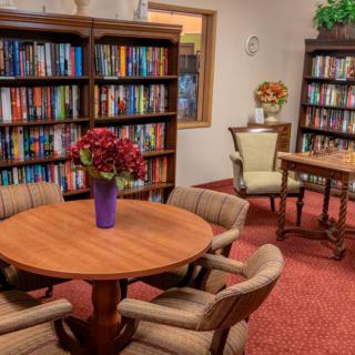 A cozy library room with bookshelves, a round table, chairs, and a vase of flowers.