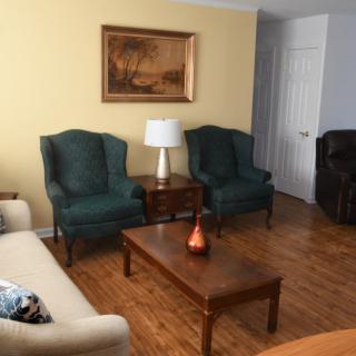 A cozy living room with a sofa, two armchairs, a coffee table, and a lamp, featuring wall artwork and hardwood flooring.