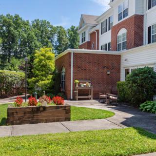 A well-maintained garden area with a brick building in the background, featuring vibrant plants and a grassy lawn.