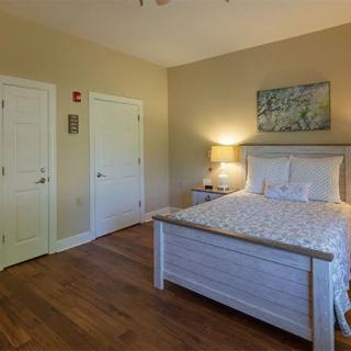 A cozy bedroom with a neatly made bed, wooden floor, nightstand with a lamp, and wall art.