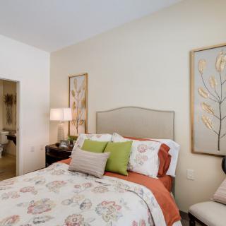 A cozy and beautifully decorated bedroom with a floral comforter, decorative pillows, artwork on the walls, and a bedside lamp.