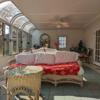 A sunlit room featuring wicker furniture and a red blanket draped over a sofa, surrounded by windows and indoor plants.