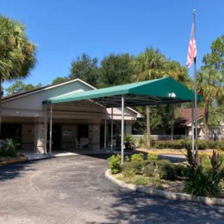 A sunny day at the entrance of a building with a green awning, surrounded by palm trees and landscaping, featuring an American flag.