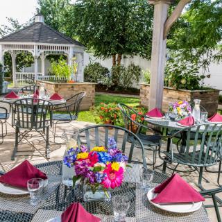 An elegant outdoor garden setting with tables adorned with flowers and burgundy napkins, surrounded by greenery and a gazebo.