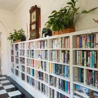 A cozy hallway library with white bookshelves filled with various books, adorned with plants and a clock, on a black and white checkered floor.