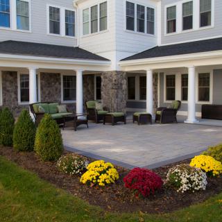 A cozy patio with outdoor seating surrounded by landscaped gardens and colorful flowers in front of a two-story house.