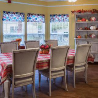 A cozy dining room with a large table set for guests, surrounded by chairs and illuminated by natural light from the windows.