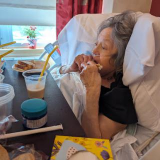 A senior person enjoying a snack in a hospital setting, surrounded by personal items.