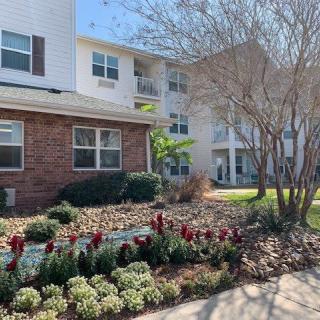 A well-maintained apartment building with landscaped gardens featuring flowers and trees, under a clear blue sky.