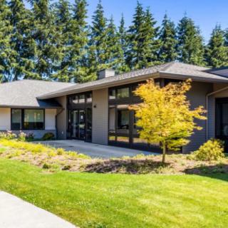 A modern, single-story house with large windows and well-maintained landscaping, surrounded by tall trees.
