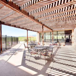 A sunlit patio with a wooden pergola casting shadows, featuring metal outdoor seating and glass doors in a modern setting.