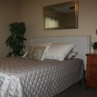 A cozy bedroom with a neatly made bed, accented by decorative pillows and a quilted comforter. A mirror hangs above the bed, and a nightstand with a lamp is placed beside it. A plant adds a touch of greenery to the space.