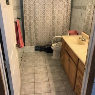 A bathroom with tiled flooring, featuring a toilet, sink with cabinets, and a shower curtain.