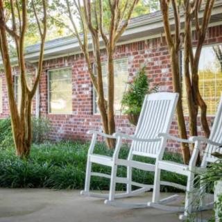 Two white rocking chairs on a patio with trees and a brick house in the background.