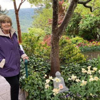 A woman enjoying a serene moment in a lush garden with blooming flowers and a Buddha statue.