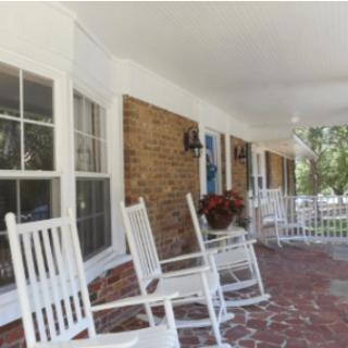 A cozy porch featuring white rocking chairs lined up against a brick wall, with windows and a floral arrangement.