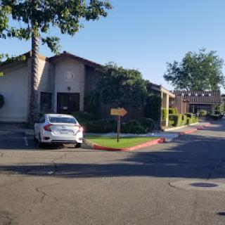 A view of a residential building with a parked car, surrounded by greenery and sunlight.