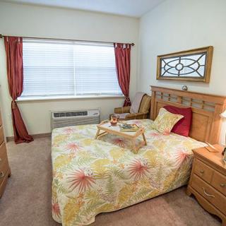 A cozy bedroom featuring a bed with floral bedding, a wooden headboard, a mirror above the headboard, and red curtains framing the window.