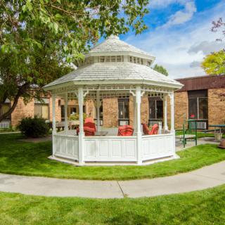 A white gazebo situated in a garden with a pathway, surrounded by trees and buildings.