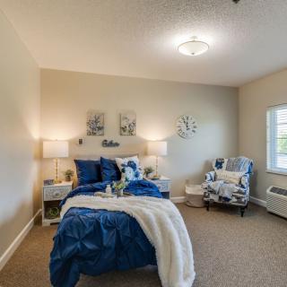 A well-decorated bedroom featuring a cozy bed with blue and white bedding, a comfortable chair, and warm lighting from lamps on either side.