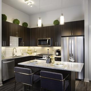 A modern kitchen with dark cabinets, stainless steel appliances, and an island countertop illuminated by pendant lighting.
