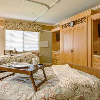 A well-furnished hospital room featuring a patient bed with a tray table, wooden wardrobe, and a window with curtains.