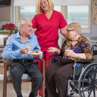 An elderly man and woman enjoying a meal with the support of a caregiver in a nursing home setting.