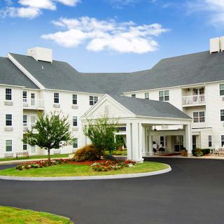 A large white residential-style building with a circular driveway and well-maintained landscaping under a partly cloudy sky.