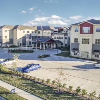 A modern apartment complex with a spacious parking lot and landscaped surroundings under a clear blue sky.