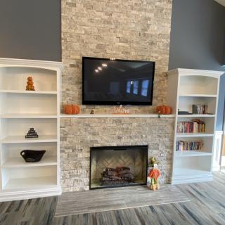 A cozy living room setup with a central stone fireplace, a mounted TV, and built-in shelves decorated with books and pumpkins.