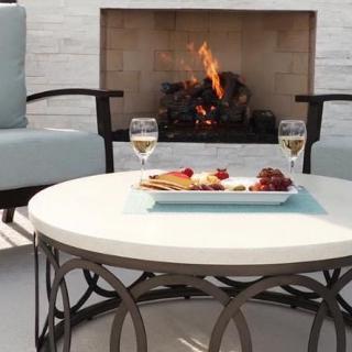 A cozy outdoor patio setup featuring a round coffee table with snacks and wine glasses in front of a warm fireplace.