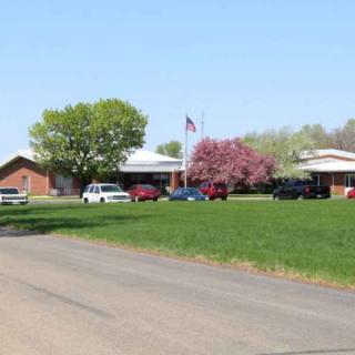 A building with a flag and cars parked in front, surrounded by trees and grass on a sunny day.