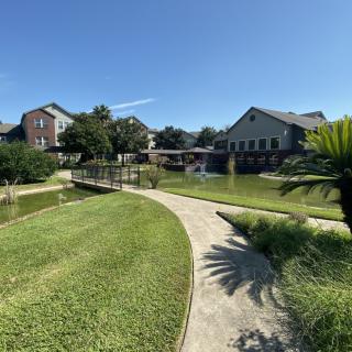 A scenic view of a landscaped garden featuring a winding pathway, pond, bridge, and several buildings under a clear blue sky.
