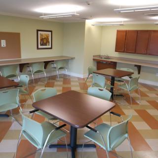 An empty break room with tables and chairs, featuring overhead lighting and wall-mounted cupboards.