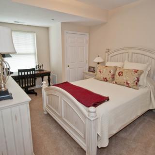 A cozy bedroom with a neatly made bed, decorated with pillows and a red throw, featuring a desk and chair in the corner.