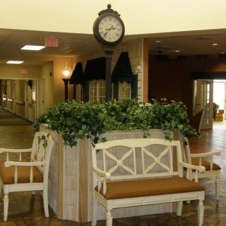 A lobby interior featuring a central seating area with benches and a decorative clock above lush plants.
