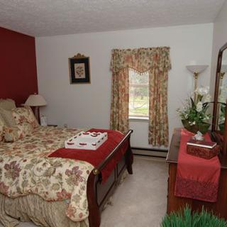 A traditional bedroom with floral patterns and warm tones, featuring a bed, dresser, lamp, and window with curtains.