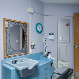 A bathroom interior featuring a blue sink, an oval mirror with decorative framing, a wall-mounted clock, cabinets, and a chair.