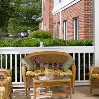 A cozy patio with wicker furniture and colorful cushions, set against a backdrop of a red brick building and lush greenery.