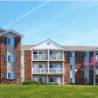 A multi-story apartment building with balconies, set against a clear blue sky and surrounded by a green lawn.