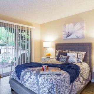 A cozy bedroom with a neatly made bed, featuring decorative pillows and a throw blanket, illuminated by a bedside lamp and natural light from large sliding glass doors.