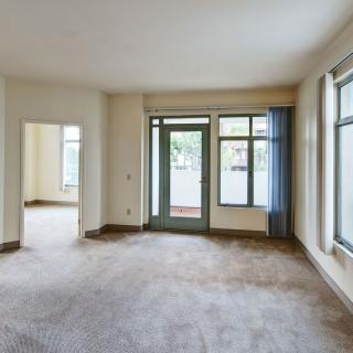 An empty living room with large windows and a glass door, featuring carpet flooring.