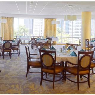 A well-lit dining room with elegantly set tables and chairs, featuring large windows with a city view.