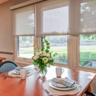 A neatly set dining table with flowers by a window on a sunny day.